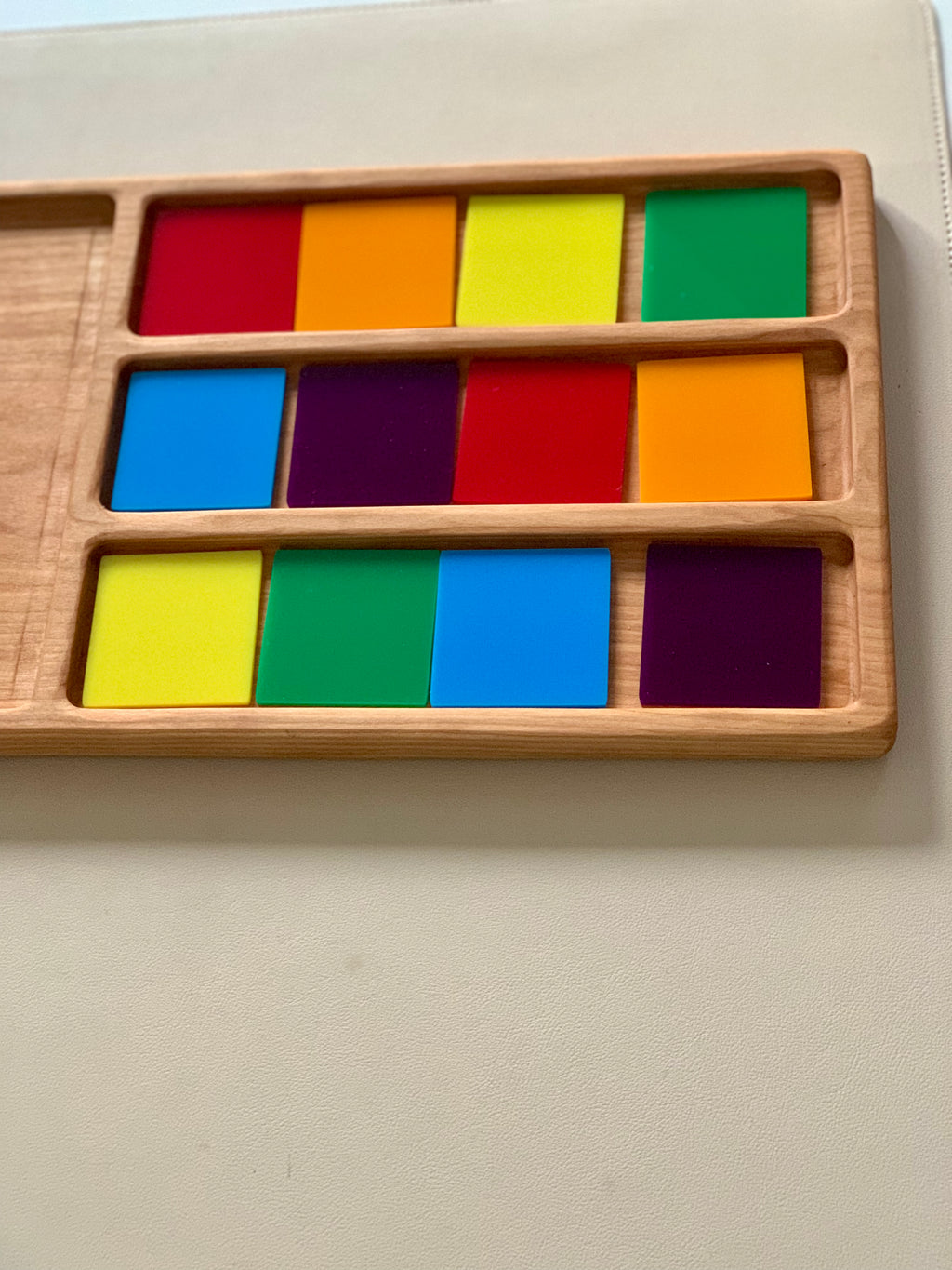 Wooden tray with three rows of colorful square tiles in red, orange, yellow, green, blue, and purple on a beige surface