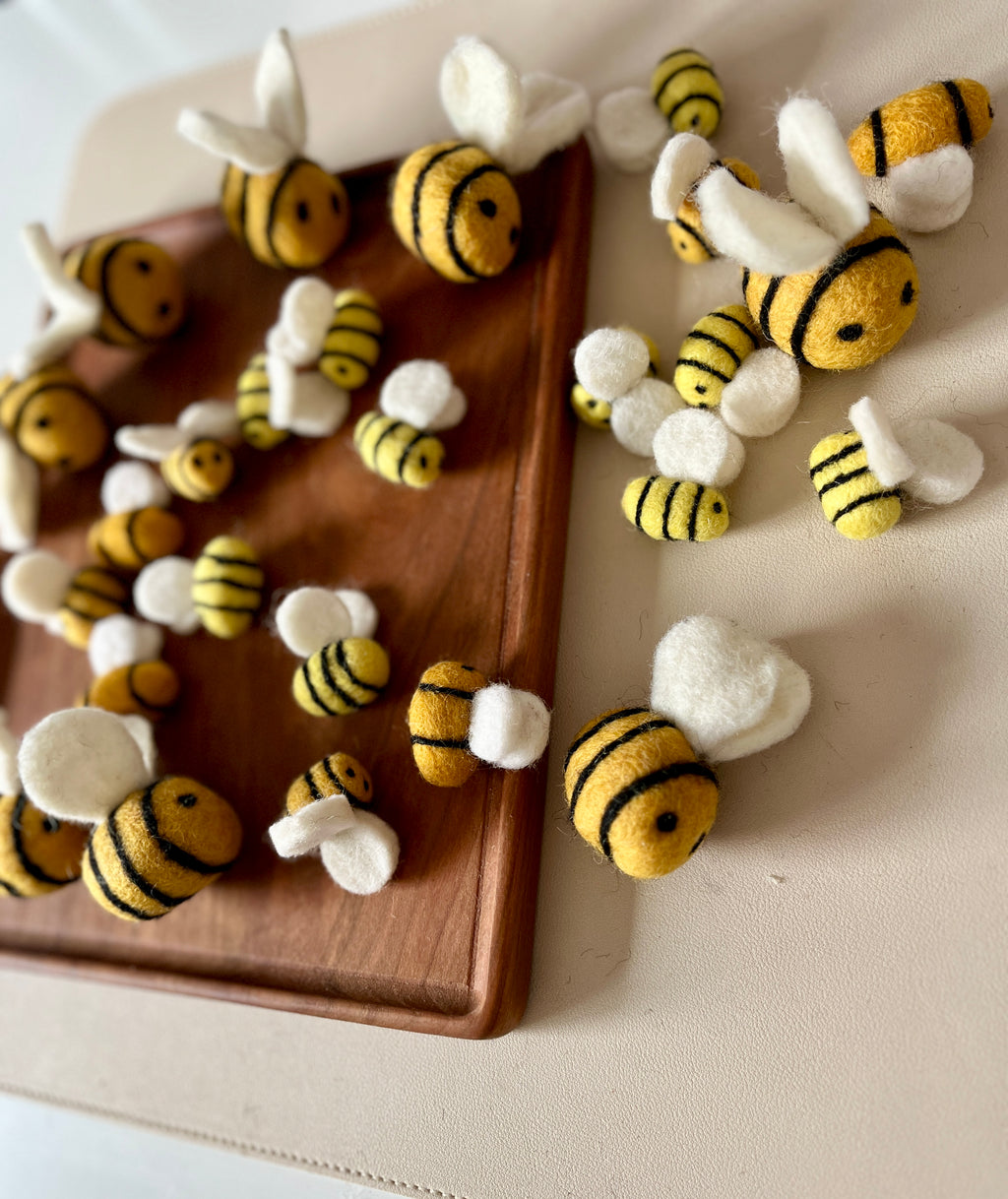 Assorted small felt bees with yellow and black stripes and white wings scattered on a wooden tray and beige surface