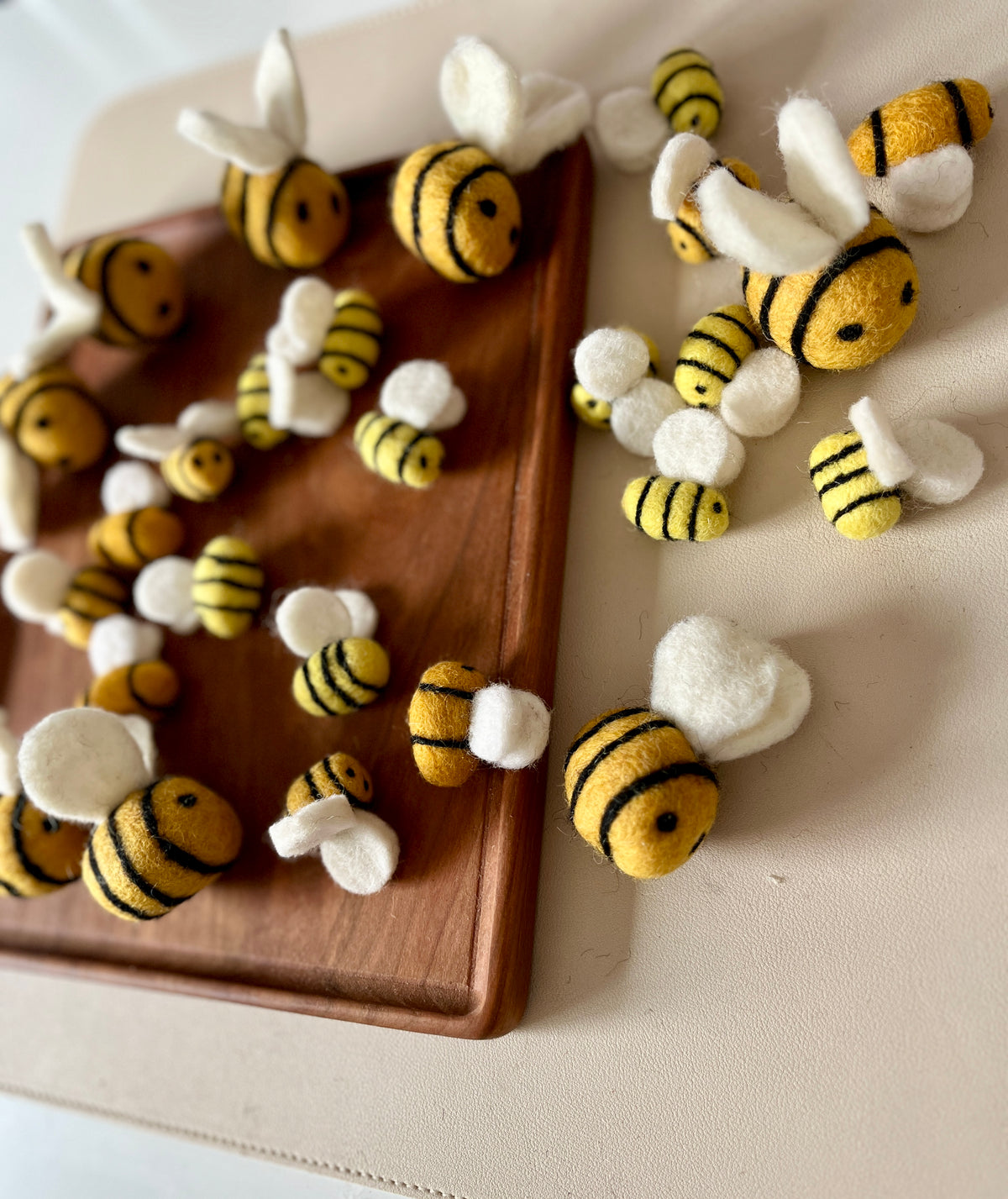 Assorted small felt bees with yellow and black stripes and white wings scattered on a wooden tray and beige surface