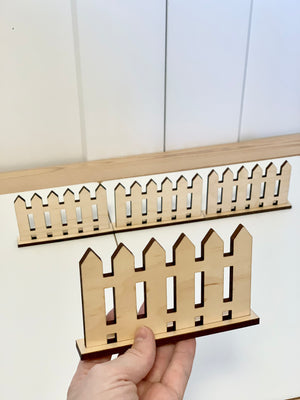 Small wooden play fences with freestanding bases arranged on white surface with hand holding one fence piece