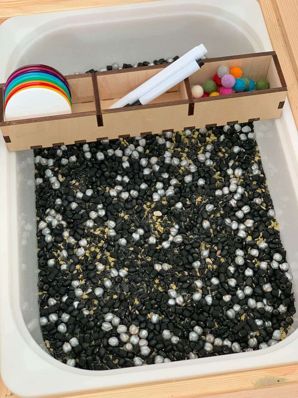 White tray filled with black and silver sensory beads, wooden divider holding markers, colorful felt balls, and round discs