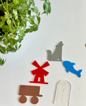 Red windmill, gray howling dog, blue fish, brown cart, and clear arched window acrylic shapes on white surface with green plant above
