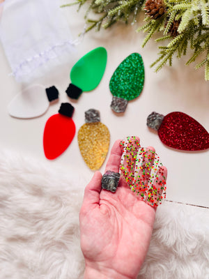 Hand holding translucent Christmas light bulb cutout with colorful confetti, surrounded by glitter and solid colored bulb shapes on soft surface