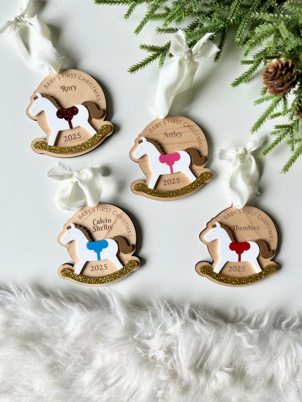 Four personalized wooden rocking horse ornaments with colorful saddles and gold glitter bases on white background with greenery