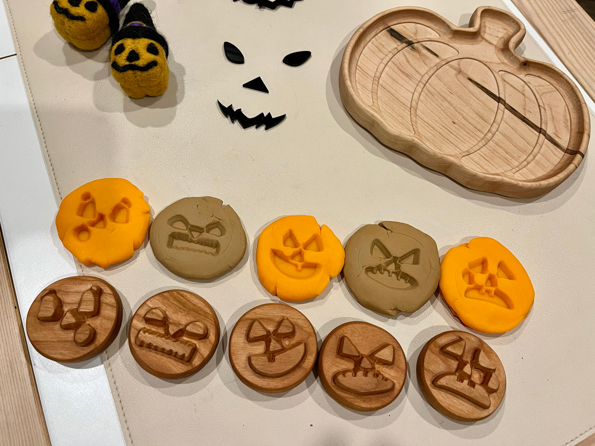 Wooden and clay discs engraved with various Halloween pumpkin faces arranged near a wooden pumpkin-shaped dish and felt pumpkin decorations