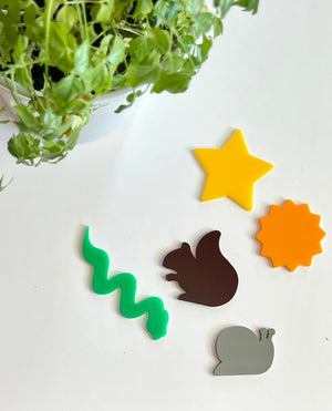 Green wavy line, brown squirrel, gray snail, yellow star, and orange sun-shaped acrylic pieces on white surface with green plant above