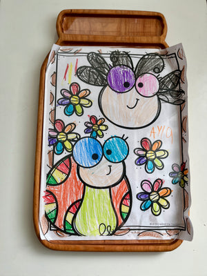 Hand-colored drawing of two cartoon bugs with large eyes surrounded by multicolored flowers on a wooden tray