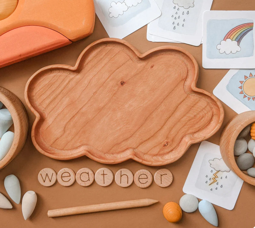 Natural wooden cloud-shaped sensory tray surrounded by weather-themed cards, wooden letters, and small toys on brown surface