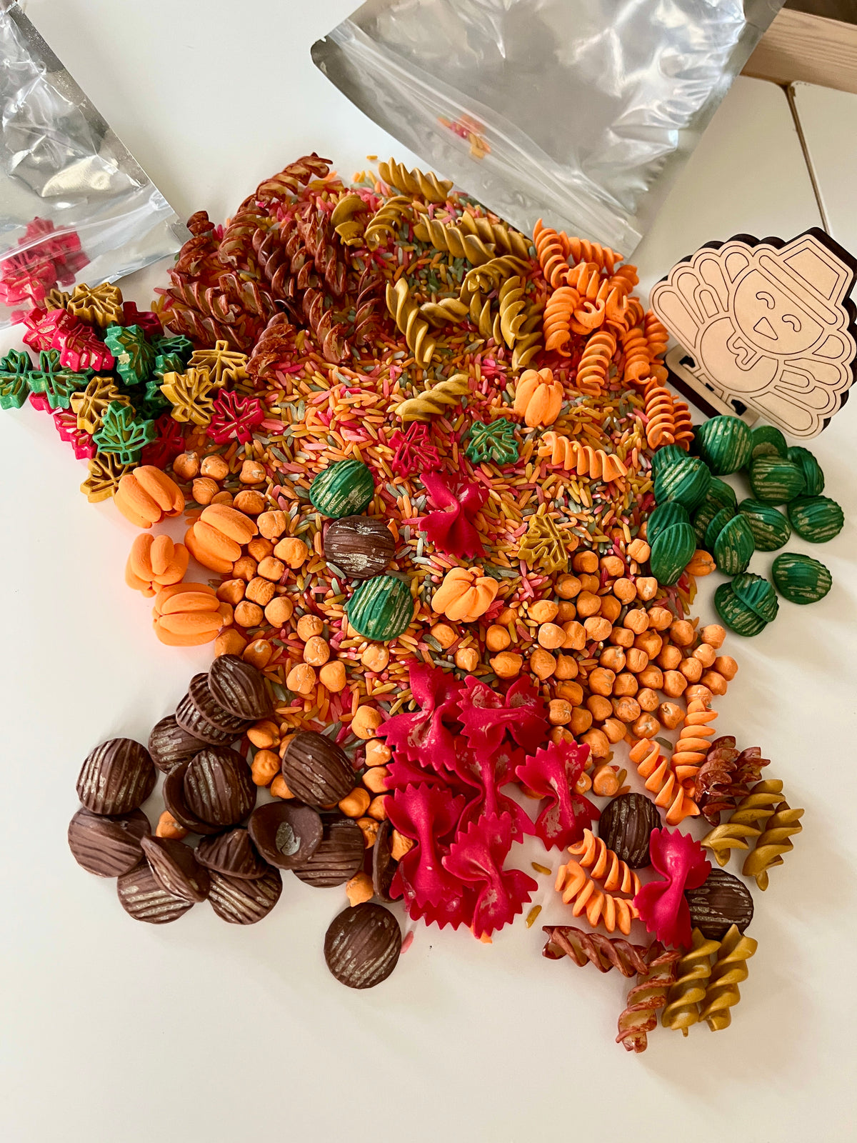 Assorted colorful fall-themed pasta and dried beans spilling from clear bags on a white surface with a wooden tag nearby