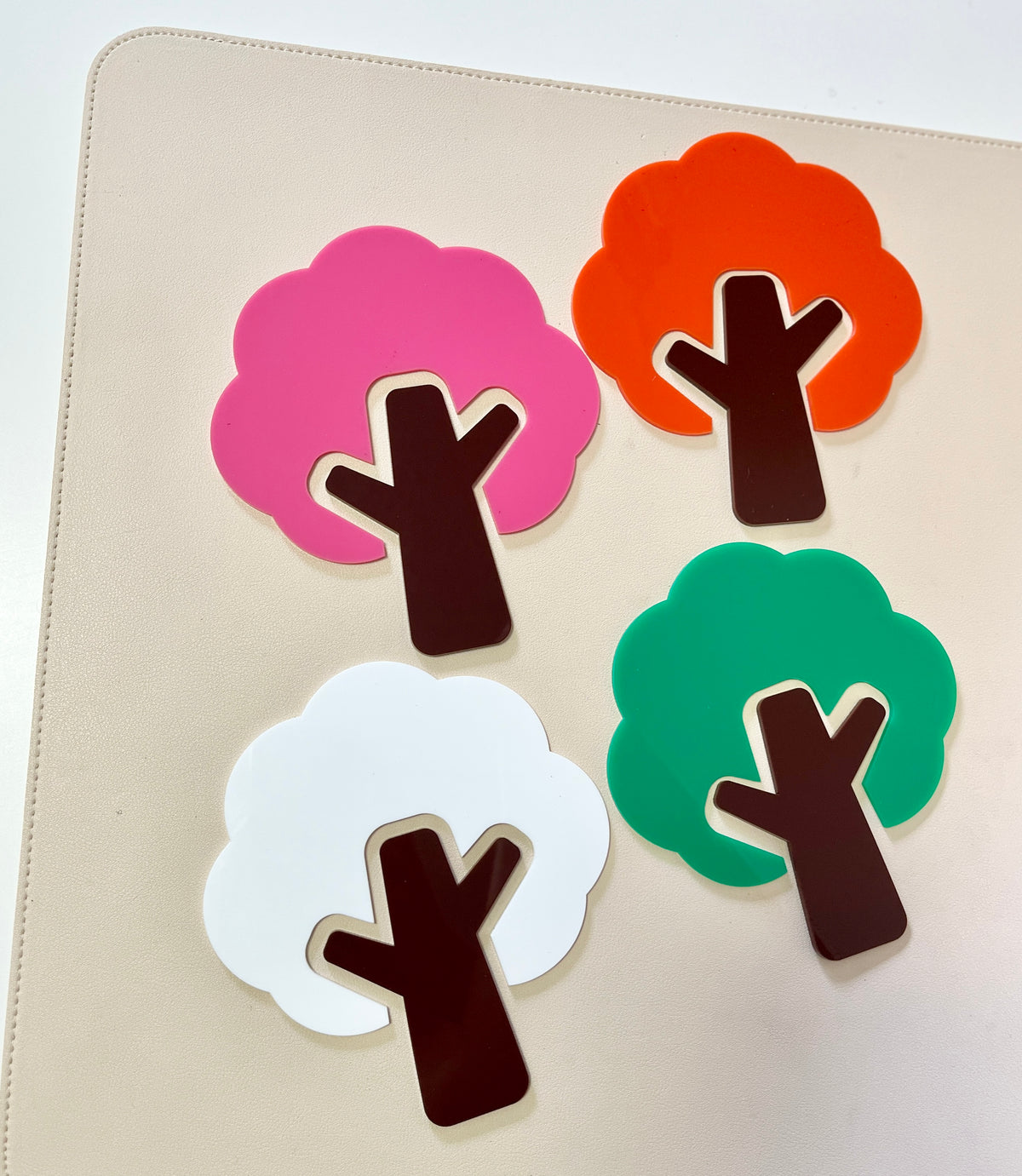 Four flat tree shapes in pink, orange, white, and green with dark brown trunks on a beige surface