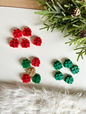Small glittery wax melts shaped like wrapped presents in red, green, and gold near pine branches on white surface