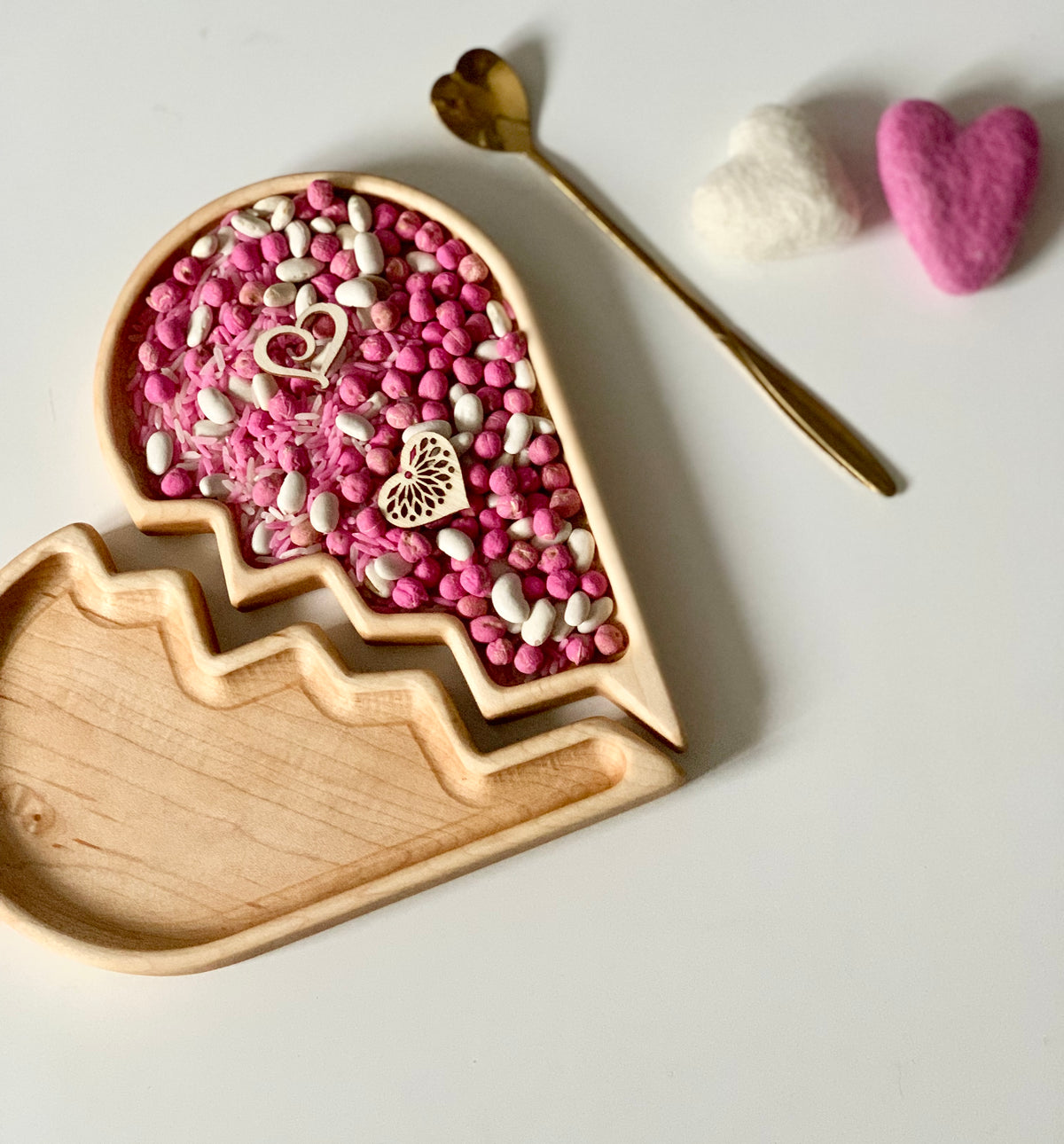 Wooden tray with heart-shaped compartments filled with pink and white beans, wooden hearts, and a gold heart-topped spoon nearby
