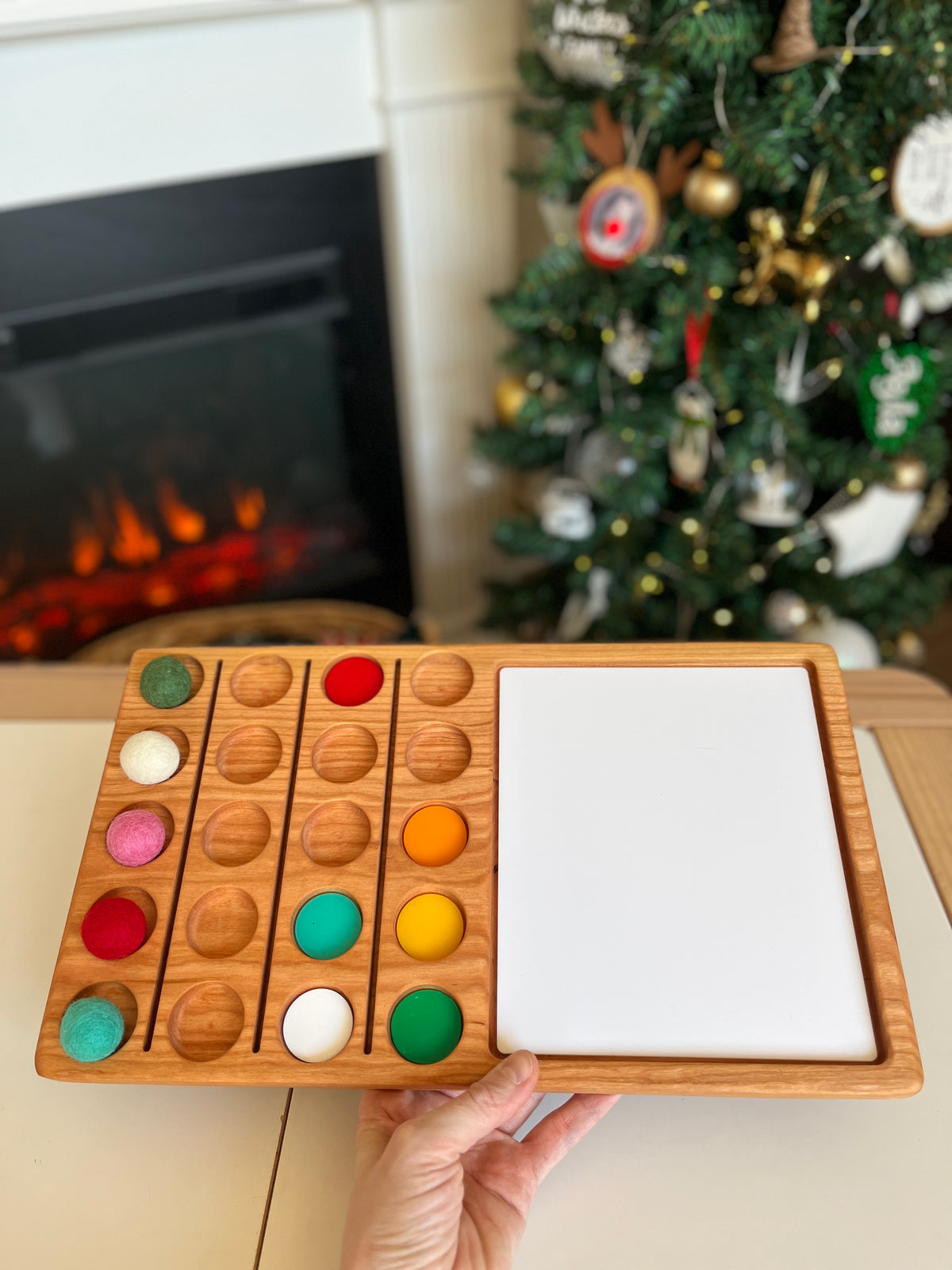 Wooden organizer with colorful felt balls in circular slots and an empty white rectangular section on a table near a Christmas tree and fireplace