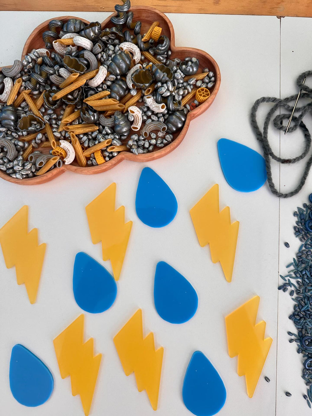 Natural wooden cloud-shaped sensory tray filled with pasta and beads, with blue raindrop and yellow lightning shapes on white surface