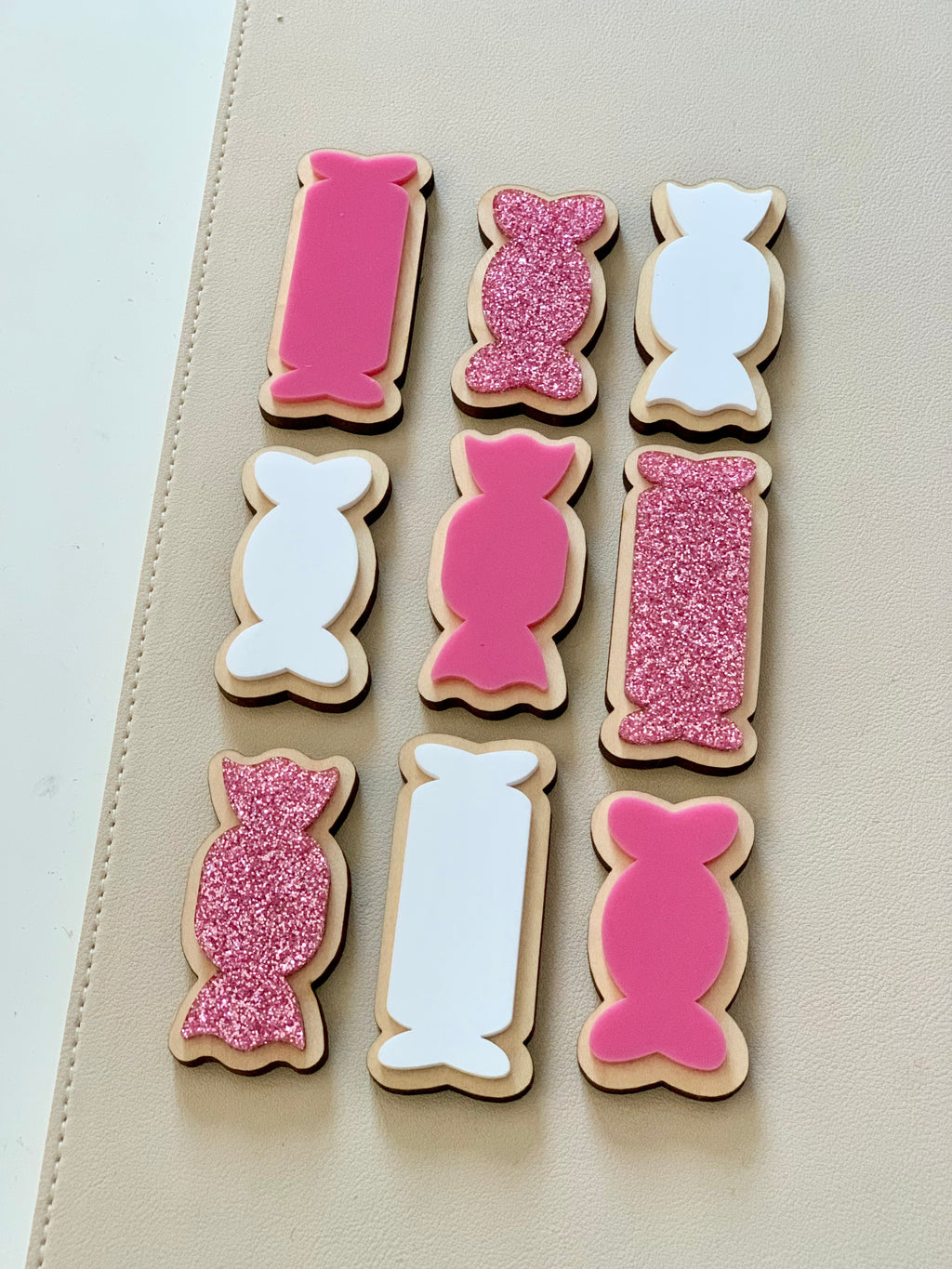 Wooden cookie-shaped pieces with pink, white, and pink glitter designs arranged on a beige surface