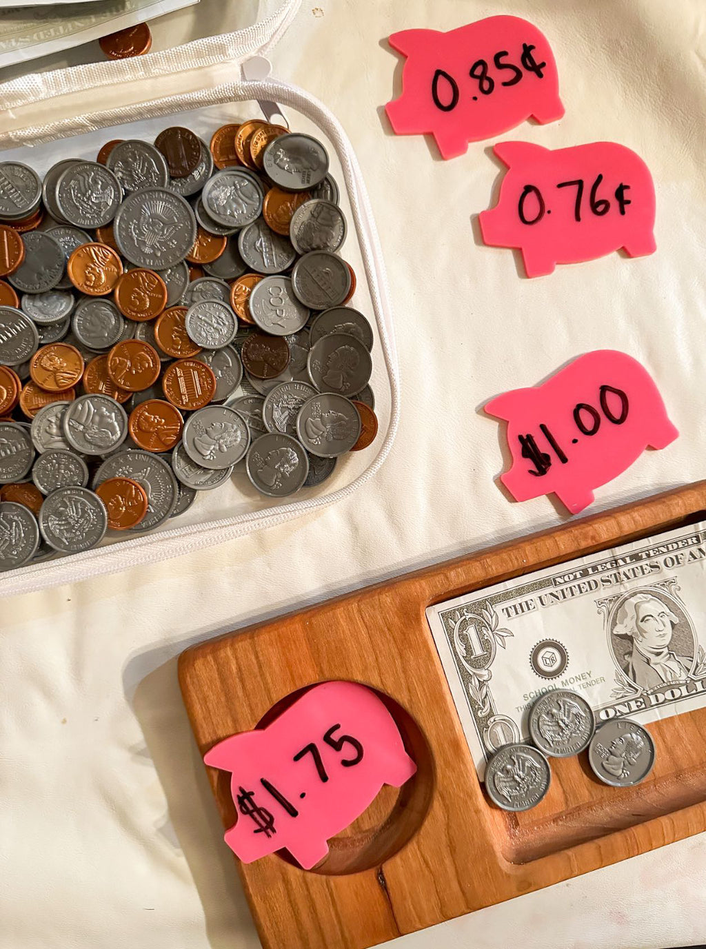 Assorted U.S. coins inside a white pouch next to pink pig-shaped price tags and wooden tray holding a dollar bill and coins