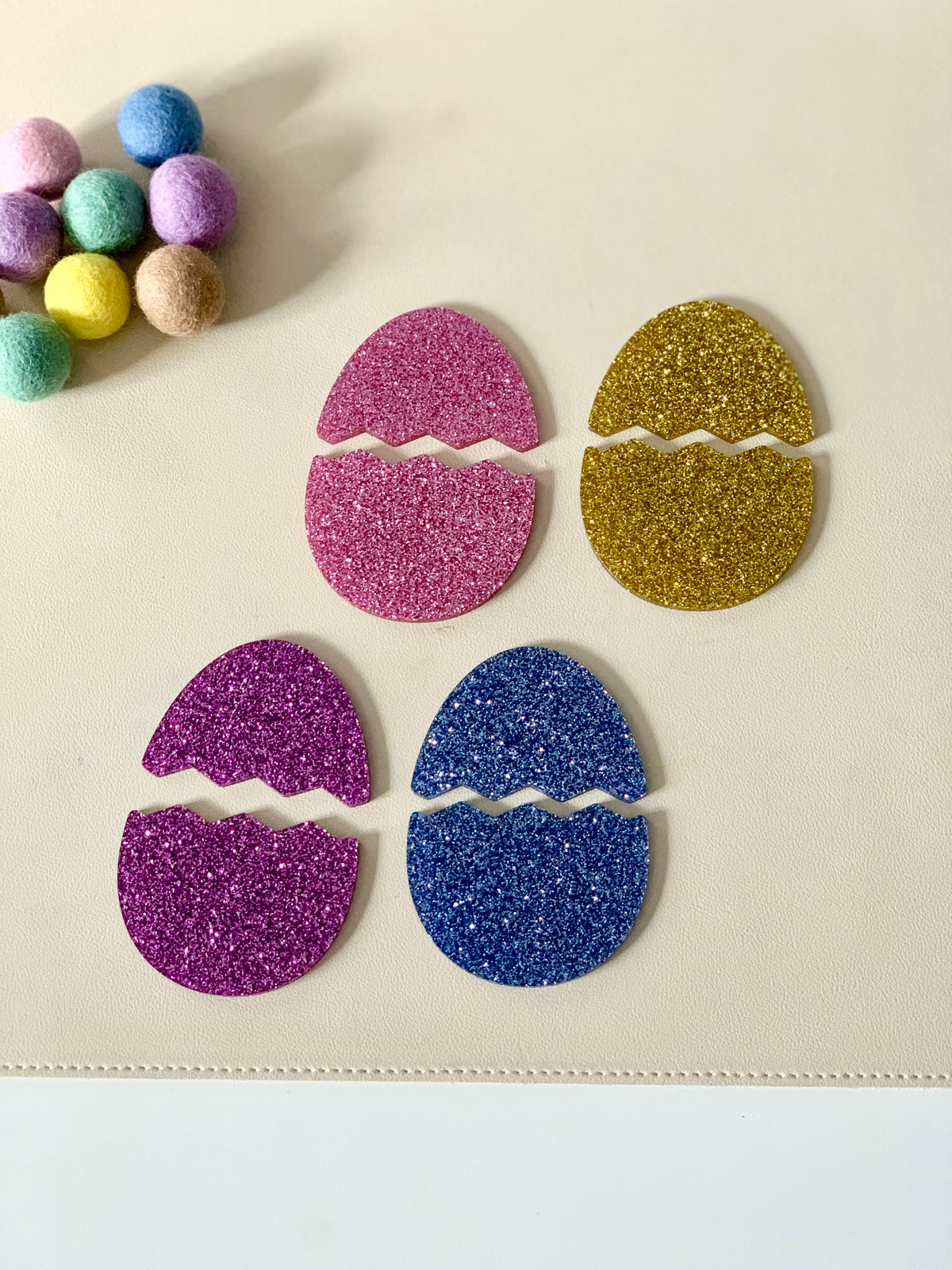 Four glittery broken egg-shaped cutouts in pink, gold, purple, and blue on a light surface near multicolored felt balls