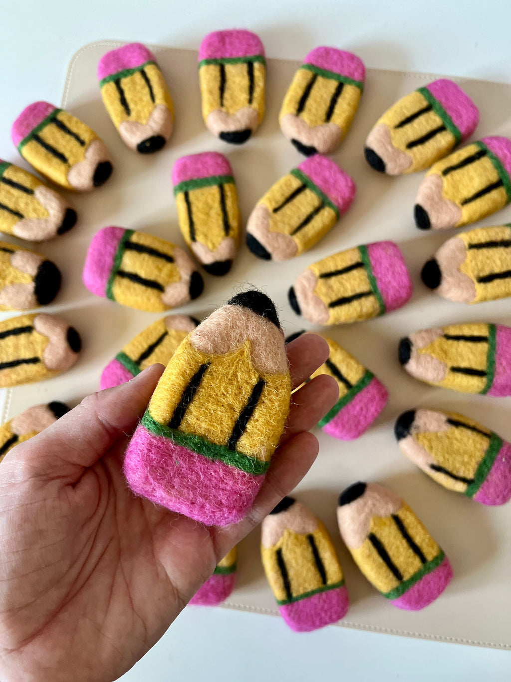 Hand holding a felt pencil-shaped toy with multiple similar toys scattered on a white surface in the background