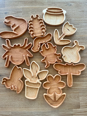 Collection of handcrafted wooden Bug Trays featuring insect and plant shapes arranged on a wood surface, designed as homeschool sensory and educational resources.