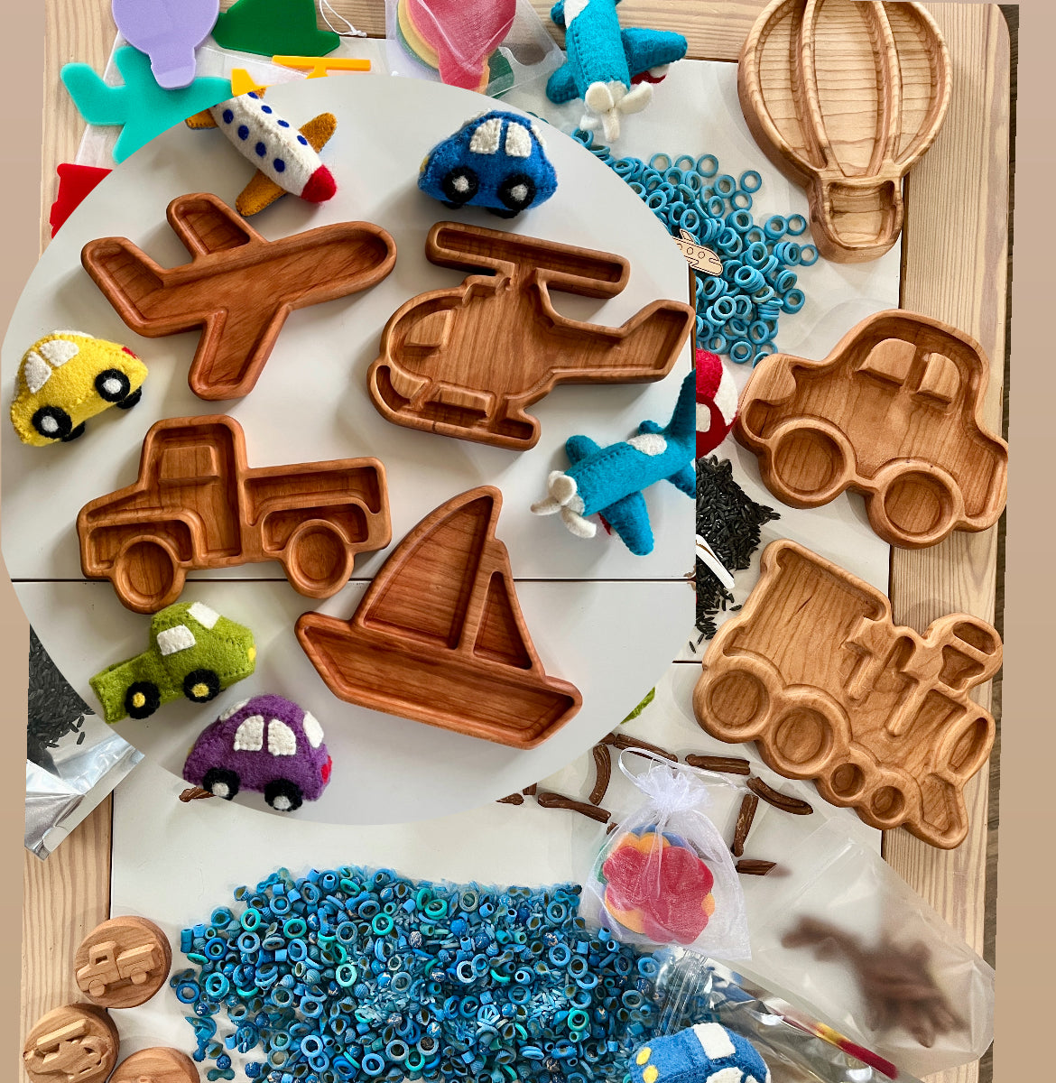 Wooden trays shaped like airplane, helicopter, car, truck, sailboat, and train with felt toys and blue pasta pieces on table