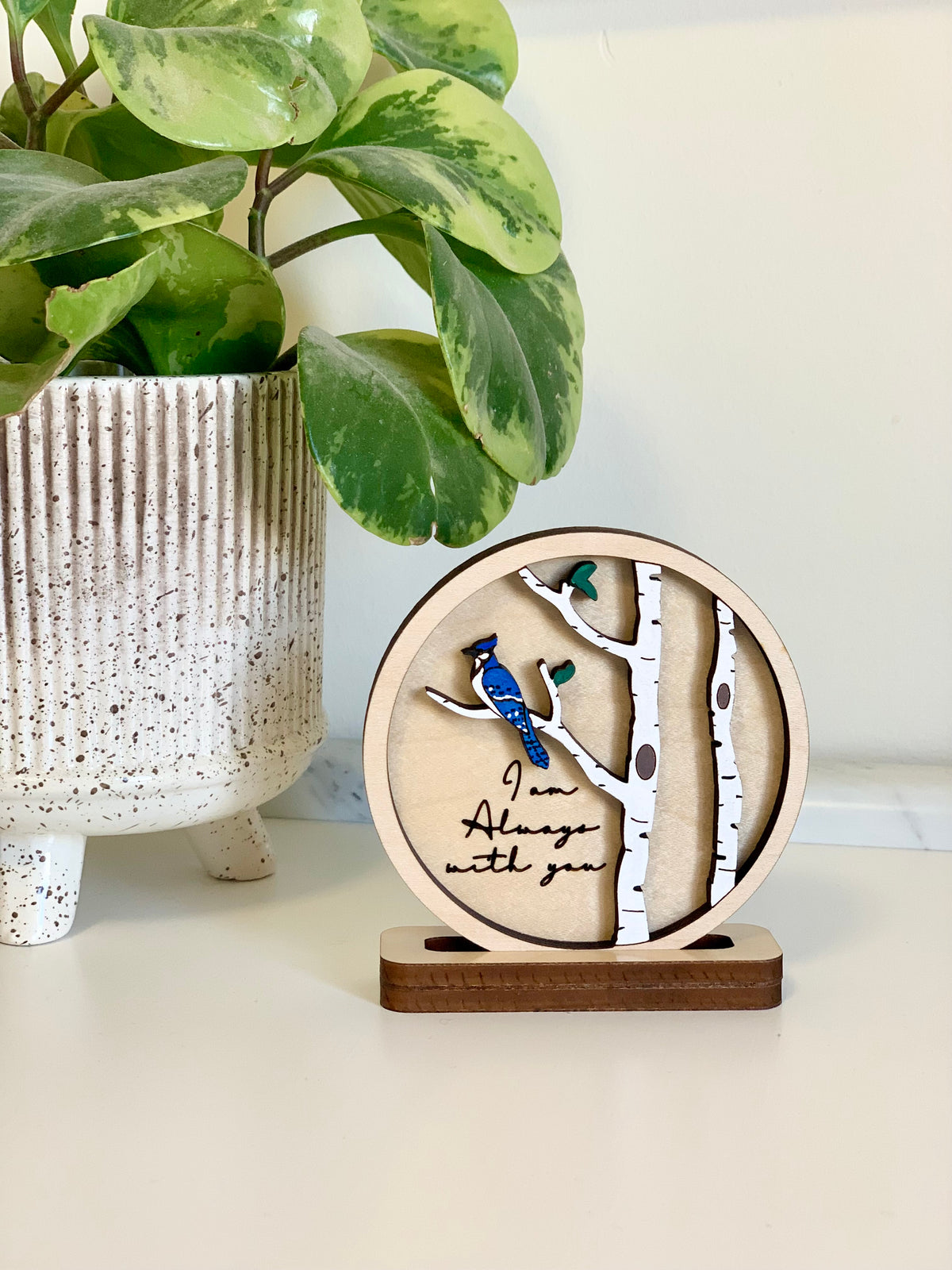 Decorative round wooden plaque featuring birch trees, a blue bird, and the phrase "I am Always with you" on a small stand next to a potted plant