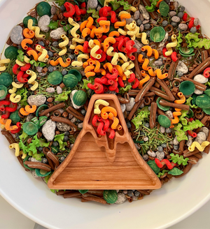 Wooden volcano-shaped dish with colorful red, orange, and yellow pasta pieces surrounded by green and brown play food items