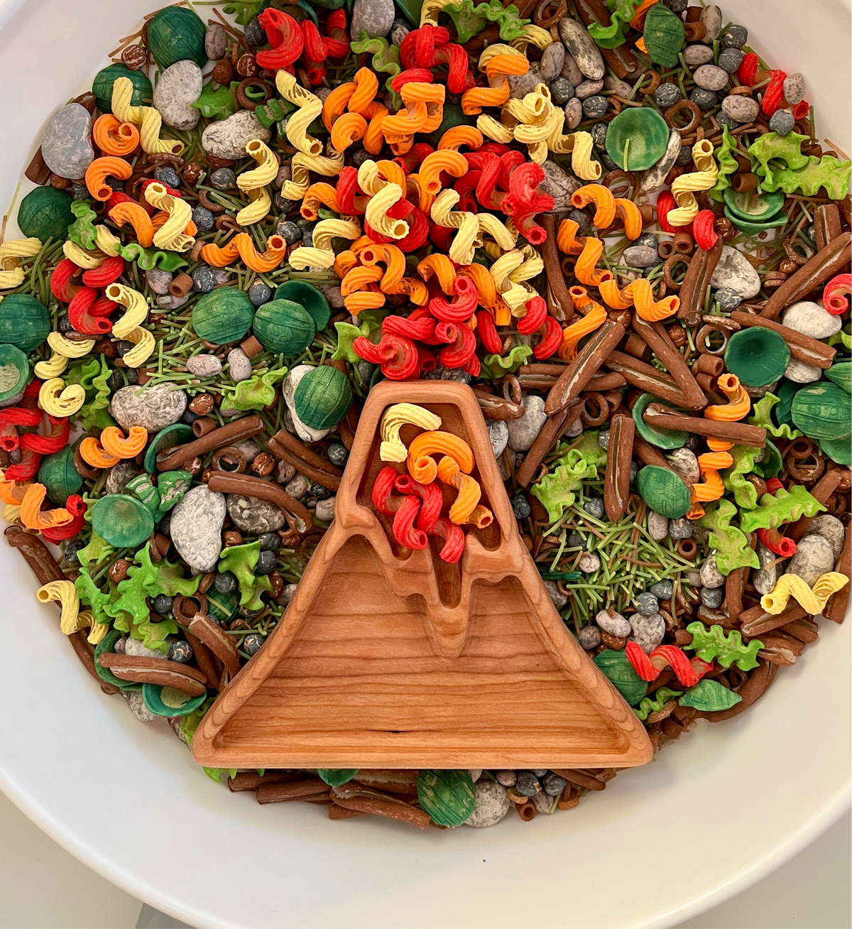 Wooden volcano-shaped dish with colorful red, orange, and yellow pasta pieces surrounded by green and brown play food items