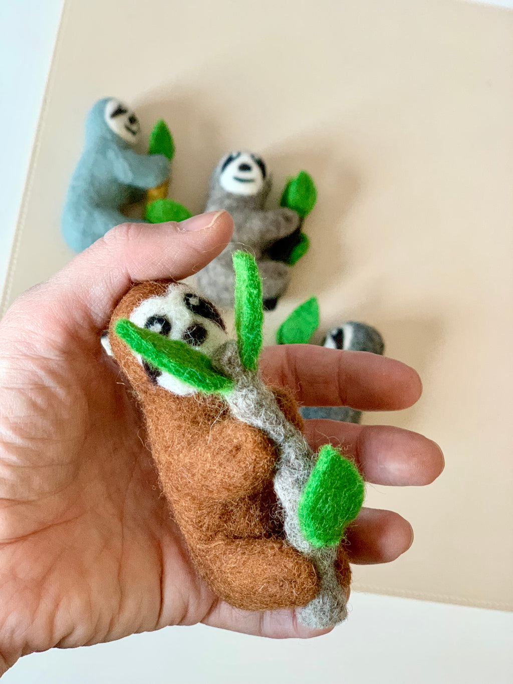 Hand holding a brown felt sloth figurine clinging to a twig with green leaves with three other felt sloths in soft colors in the background