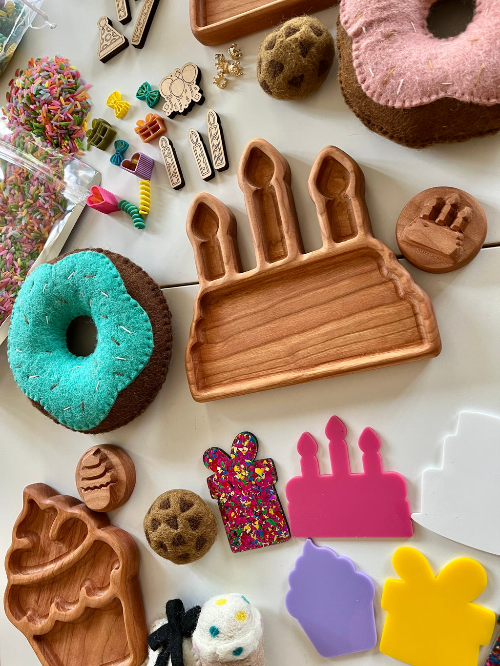 Wooden birthday cake tray and candle stamp surrounded by felt donuts, colorful sprinkles, cookie shapes, and crafting pieces on white surface