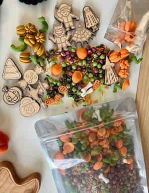 Halloween-themed sensory bin materials including wooden characters, pumpkin shapes, bones, and colorful dried pasta on a white surface
