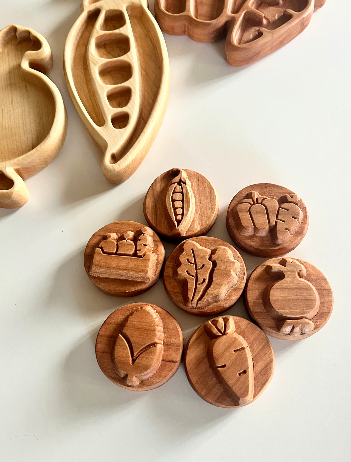 Round wooden tokens engraved with vegetable shapes arranged on a white surface near carved wooden trays