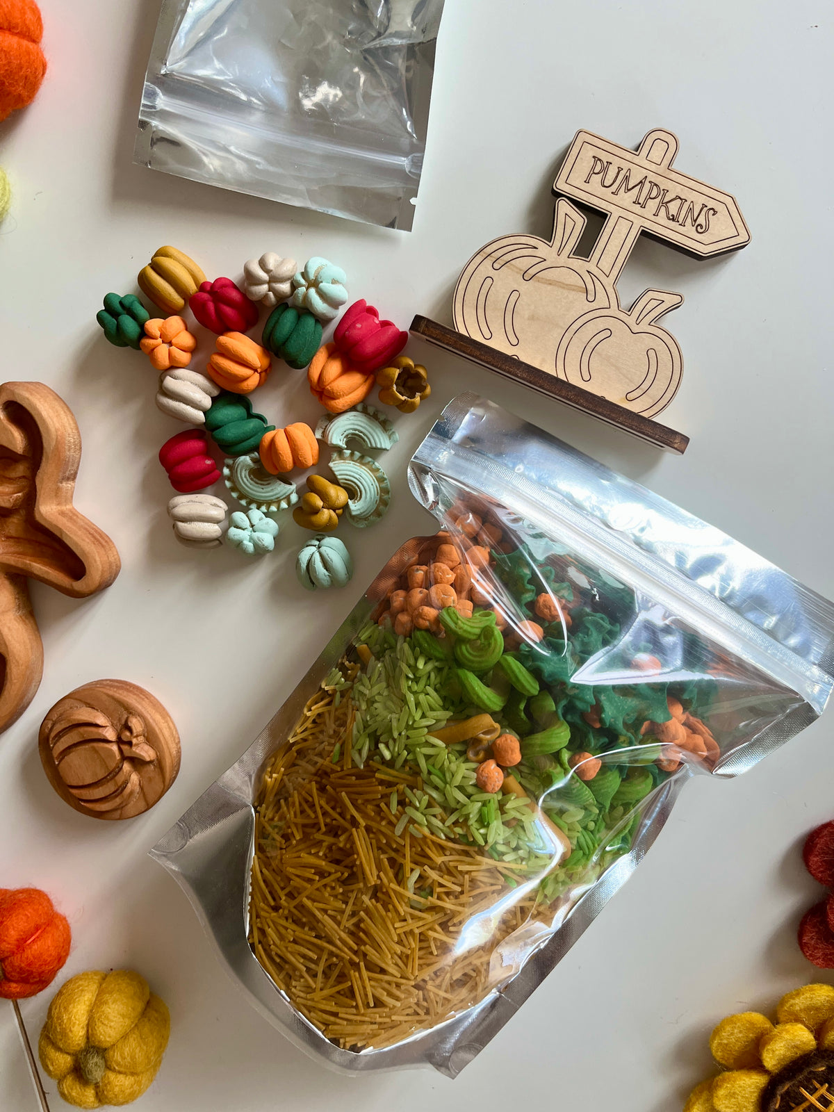 Assorted colorful pumpkin-shaped pasta pieces, small pumpkins, and a wooden "pumpkins" sign on a white surface with packaged sensory items
