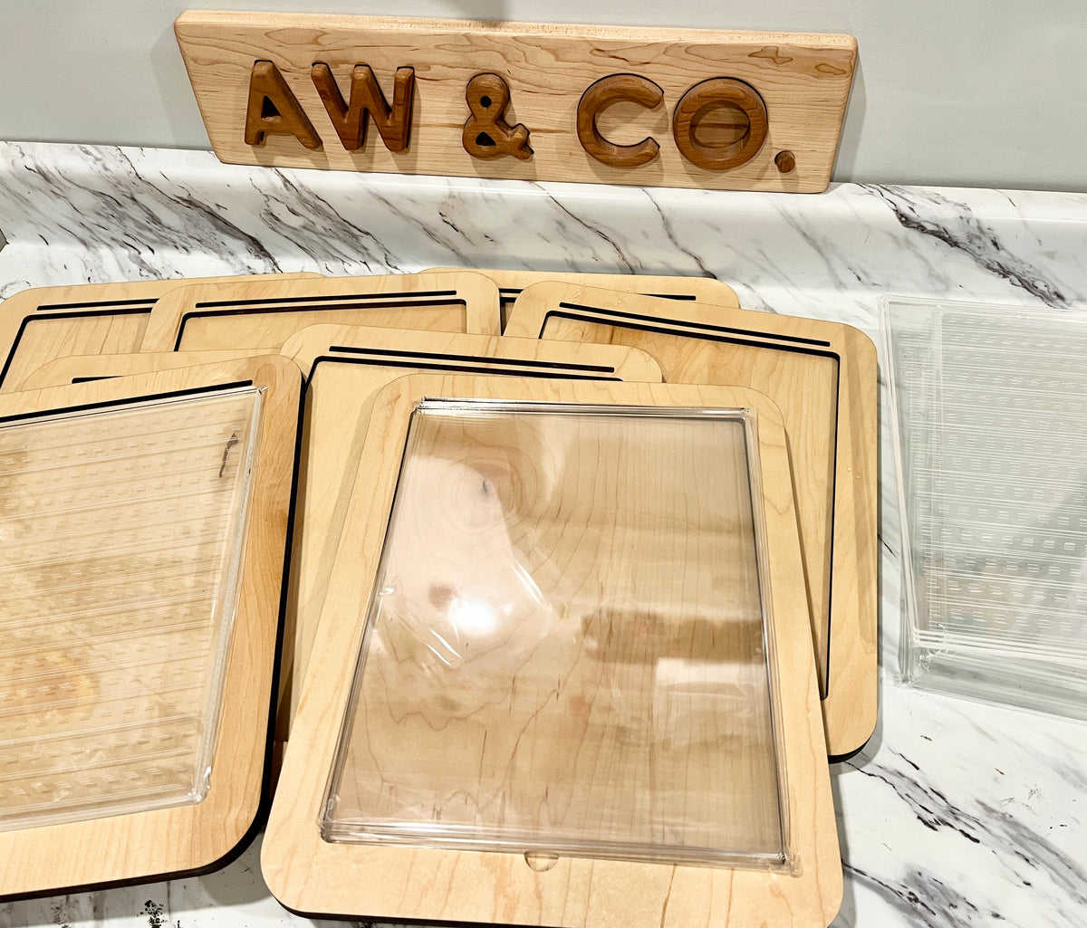Stack of wooden trays with clear plastic lids on a marble countertop below a wooden sign reading AW & CO.