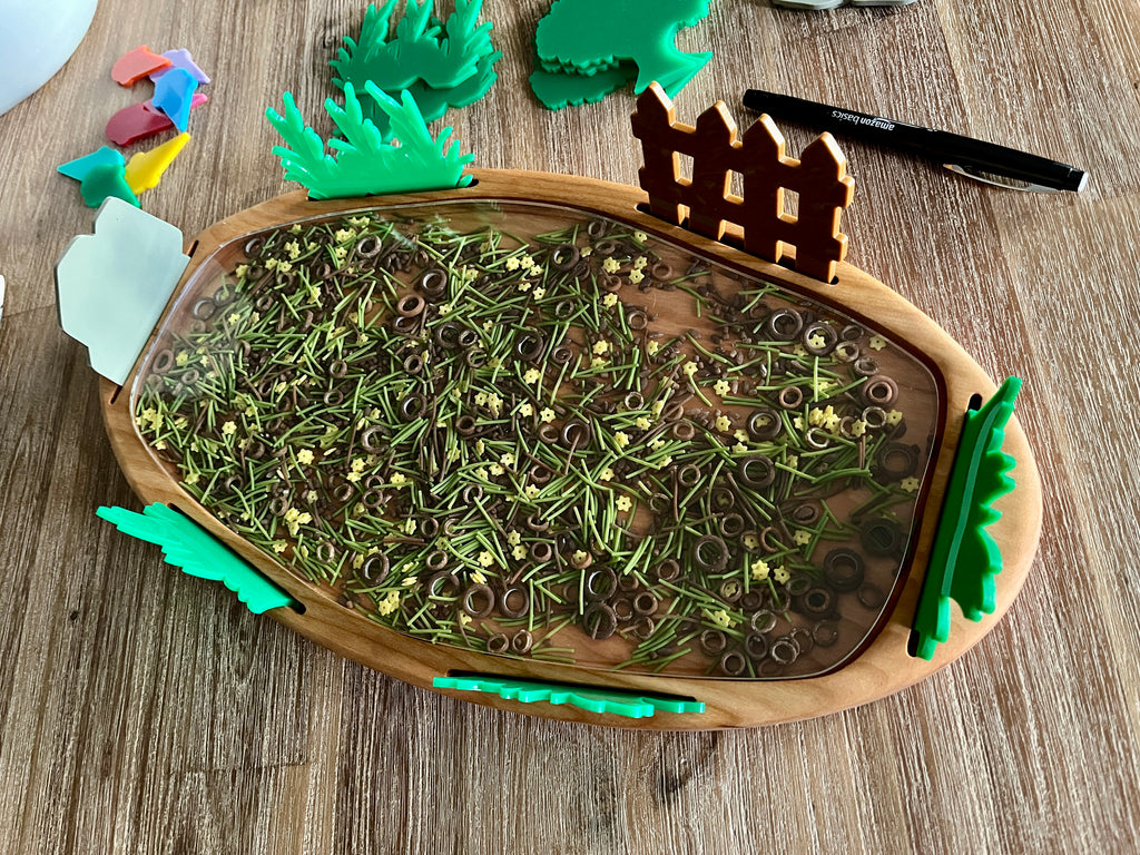 Multipurpose Oval Sensory Tray with wood and metal sensory filler, green plant-shaped accents, and wooden fence piece, designed for handcrafted homeschool education resources.