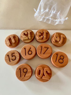 Set of round wooden tokens numbered 11 to 20 with engraved and raised digits beside a sheer white drawstring pouch