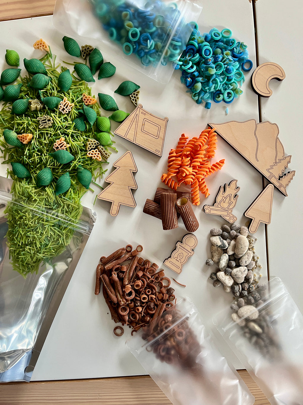 Natural and colorful textured play materials including wooden camping shapes, pasta, rocks, and plastic rings on white surface