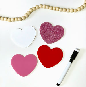 Four heart-shaped boards in white, pink, red, and glitter pink arranged with a dry erase marker and wooden beads above