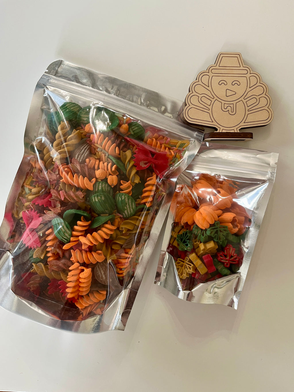 Clear bags containing spiral and shaped pasta in autumn colors next to a wooden turkey cookie cutter on white surface