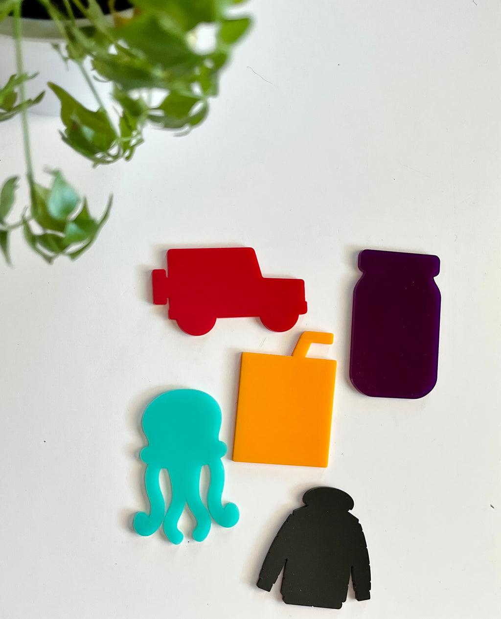 Five vibrant acrylic shapes including red jeep, purple jar, teal octopus, orange juice box, and black jacket on white background with green plant above