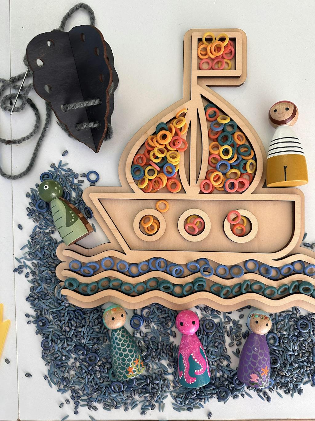 Wooden fillable boat play tray with colorful rings, wooden sea creature and people peg dolls, and blue rice grains on white surface
