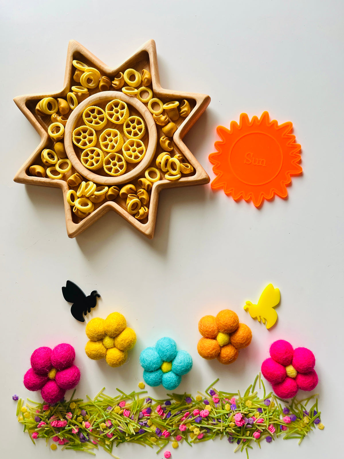 Wooden star-shaped tray with yellow pasta wheels, orange sun-shaped cutter, colorful felt flowers, bees, and green floral grass arrangement on white background