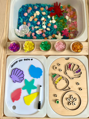 Wooden activity board with shell shapes filled with colorful translucent beads and small bowls of sorted beads above.