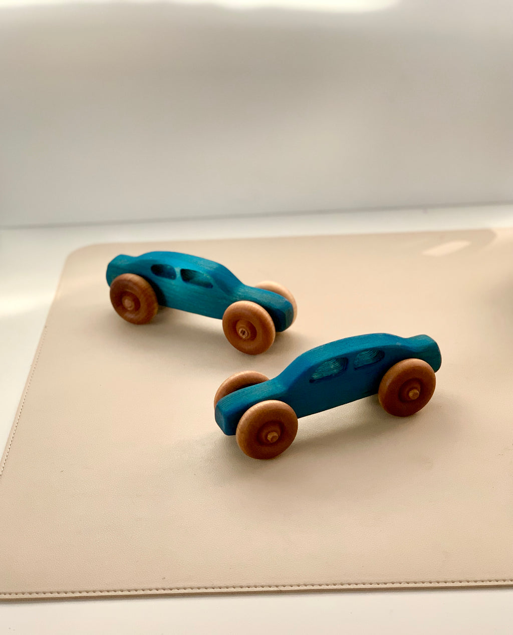 Two small blue wooden toy cars with natural wood wheels on a smooth beige surface in soft light