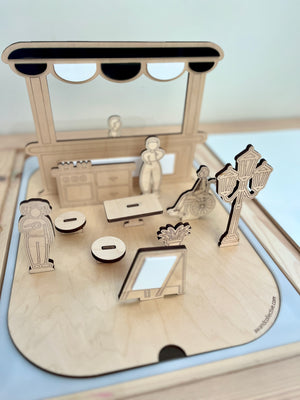 Miniature wooden scene with laser-cut figures, streetlamp, furniture, and storefront facade on a light wood base