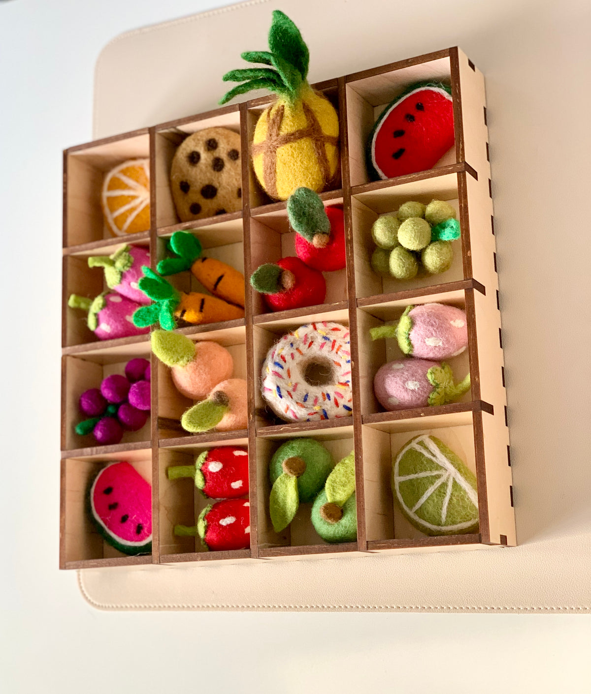 Wooden grid displaying colorful felt fruits, vegetables, and a sprinkled donut arranged in separate compartments
