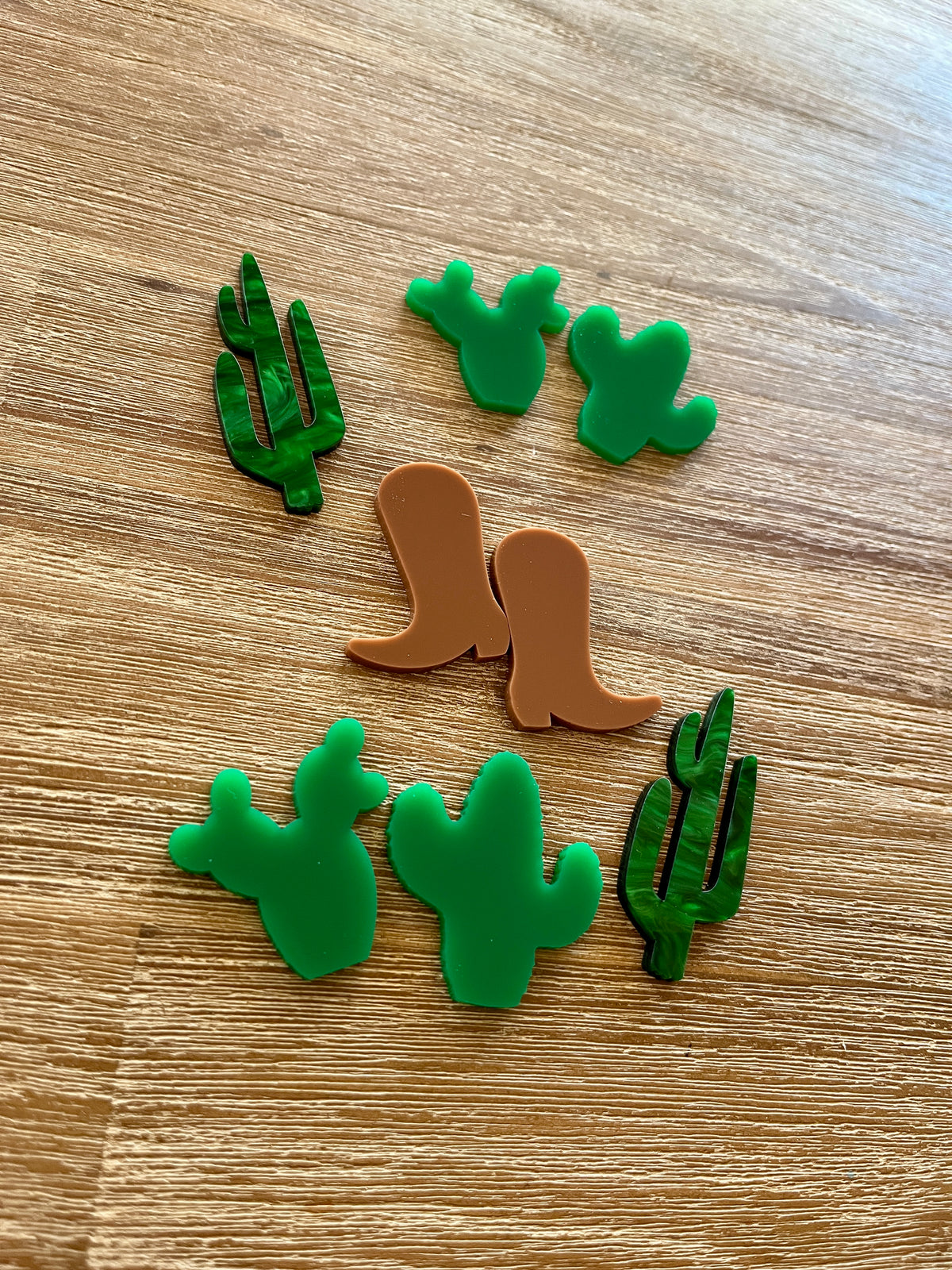 Seven small flat shapes made of durable material arranged on a wooden surface including two translucent green cacti, four solid green cacti, and a pair of brown boots, Little Dry Erasables - Cacti & Boots, handcrafted, homeschool, education resources.