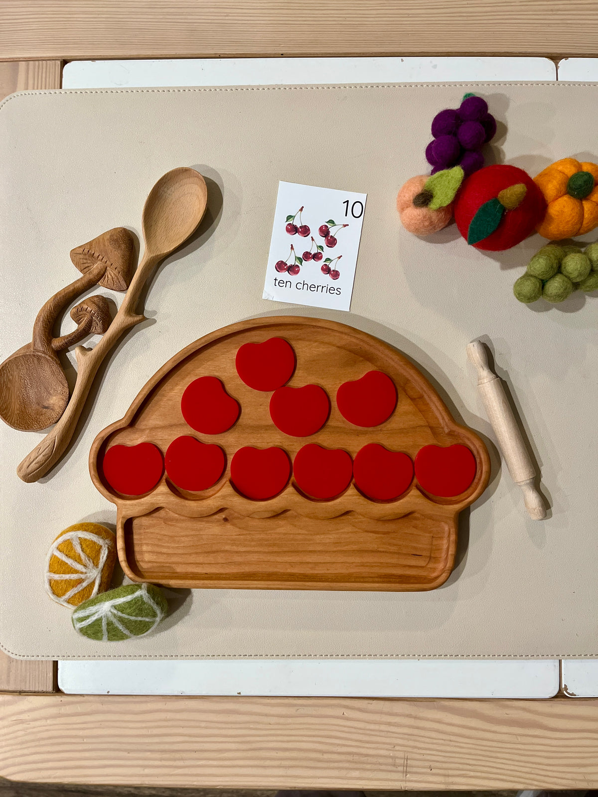 Wooden pie-shaped tray with ten red apple-shaped pieces, wooden spoons, rolling pin, felt fruits, and a flashcard on beige surface
