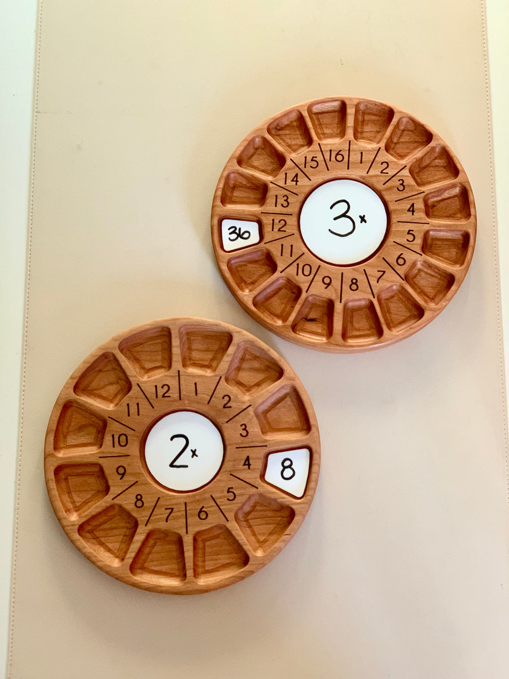 Two circular wooden multiplication learning boards with numbered compartments and central multiplier labels on a beige surface