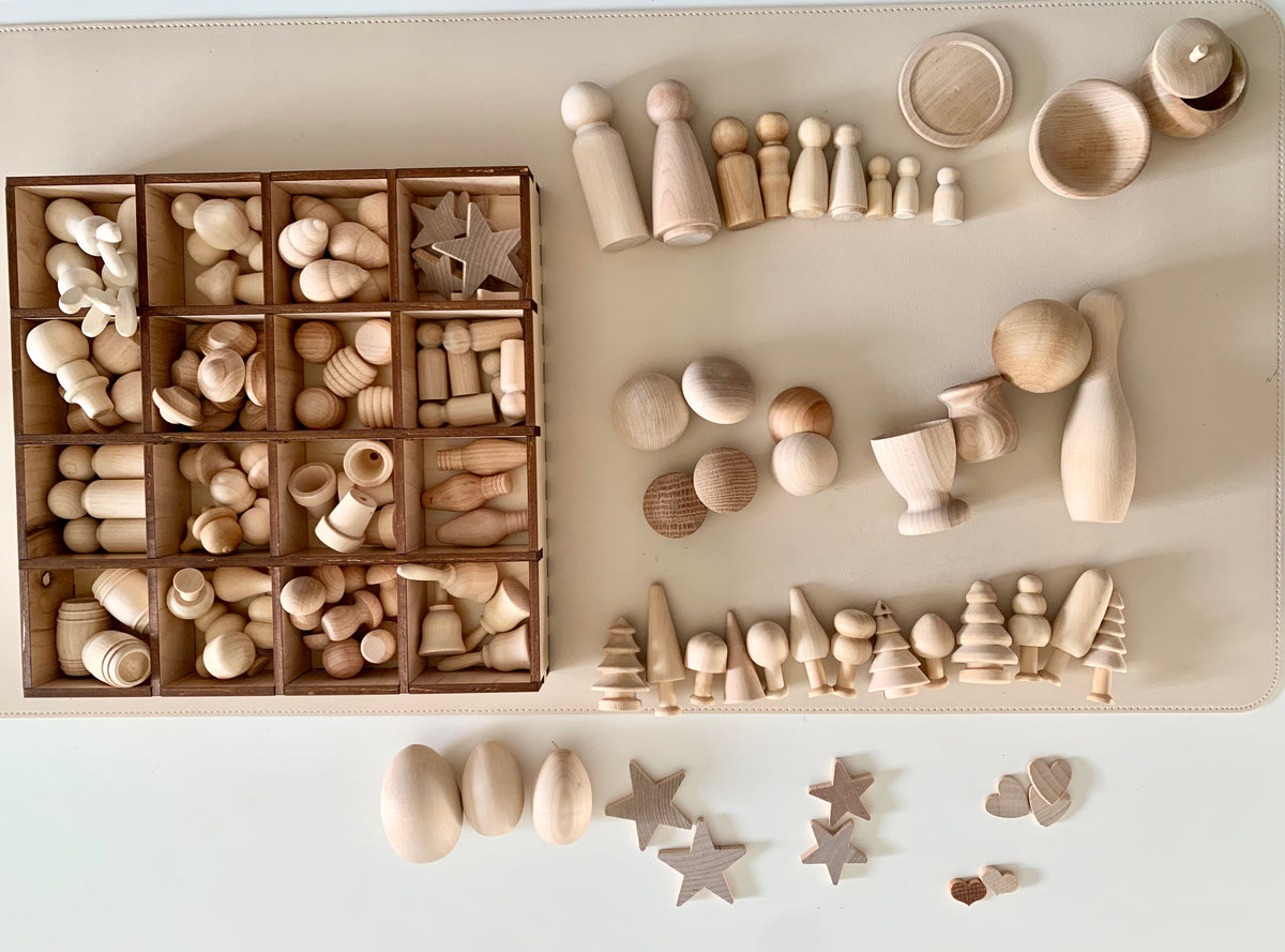 Various unpainted wooden peg dolls, balls, trees, stars, eggs, and other shapes arranged on a beige surface and in a compartmentalized tray