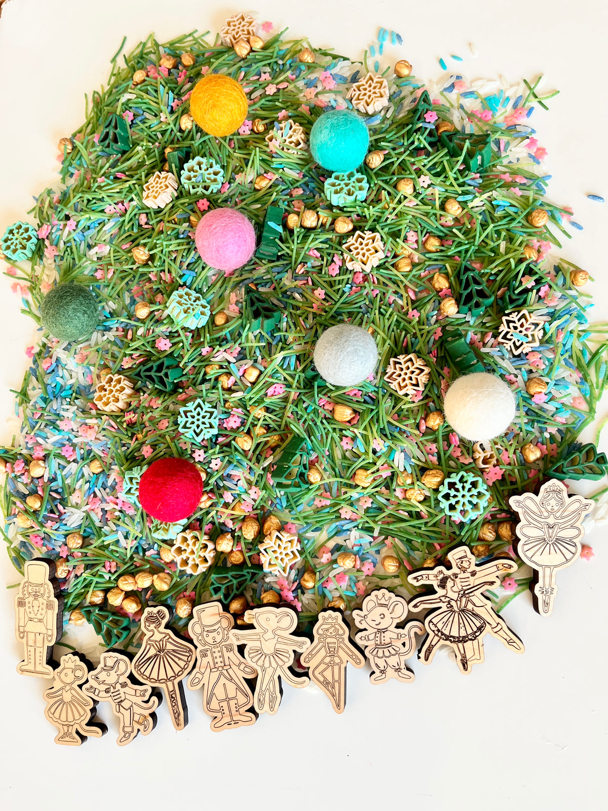 Colorful sensory mix with green pine needle pieces, felt balls, gold beads, wooden snowflakes, and ballet-themed figurines on white surface